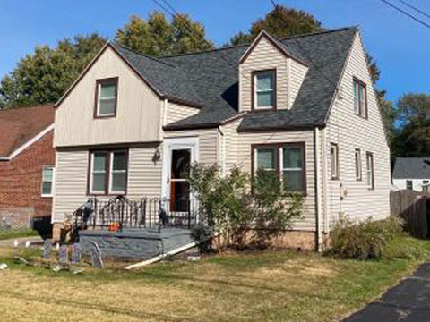 A two-story house with beige siding and a gray roof. The house has a small front yard with a few plants.