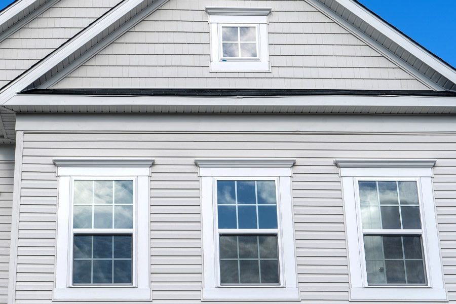 A house with three white framed windows and gray siding. The windows reflect a blue sky.
