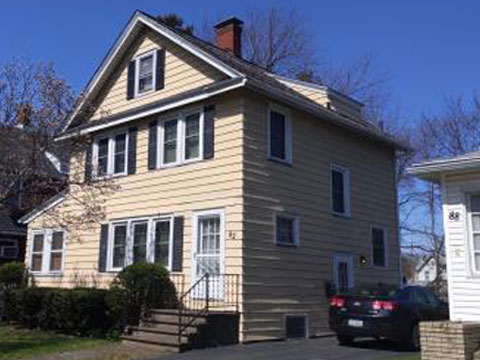 A two-story yellow house with black shutters. A black car is parked in the driveway.