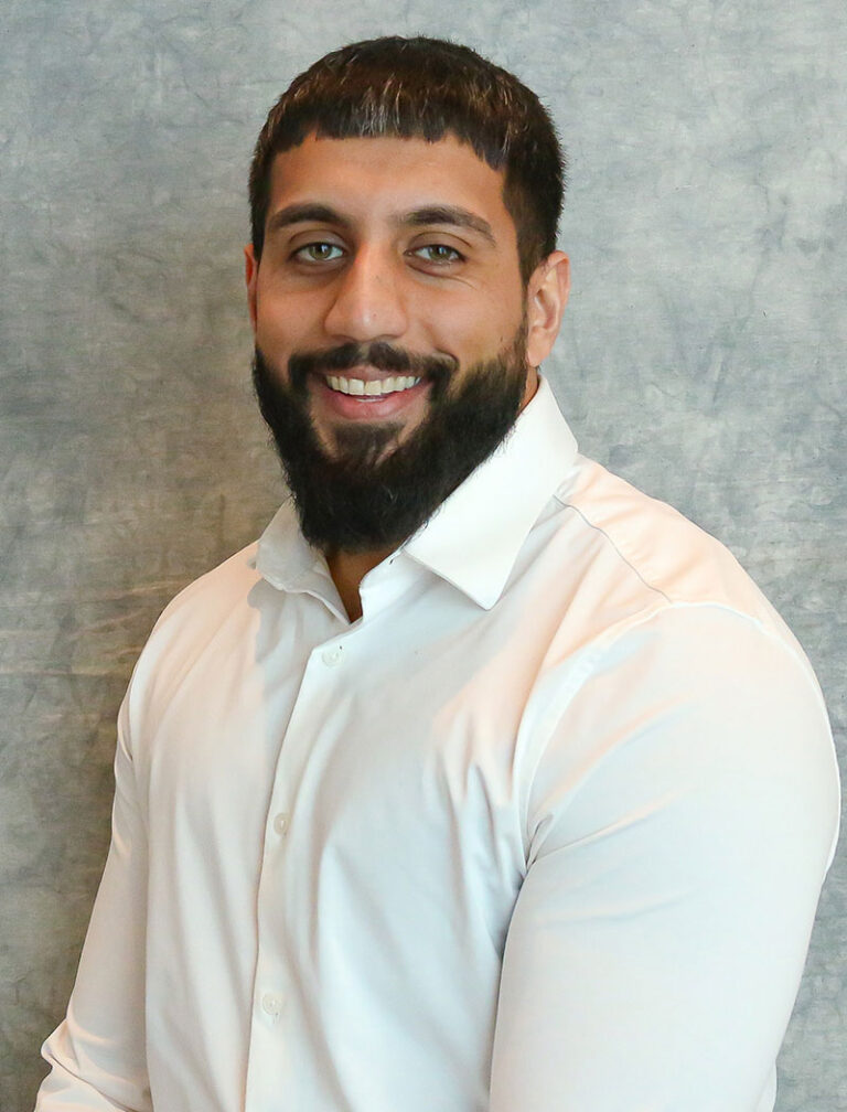 A smiling man with a beard wearing a white shirt. He is standing in front of a gray background.