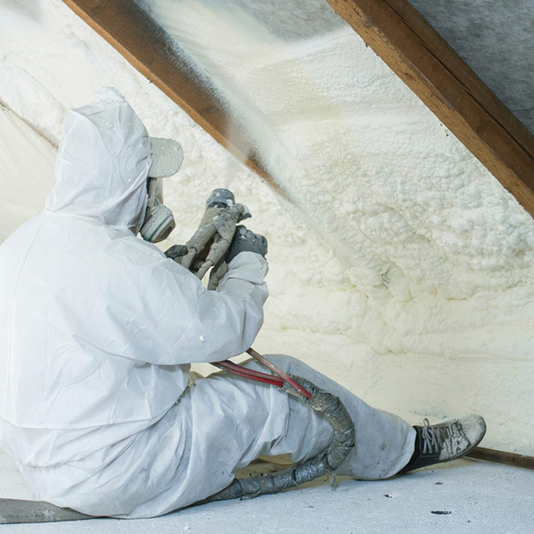 A worker in protective gear spraying insulation in an attic.