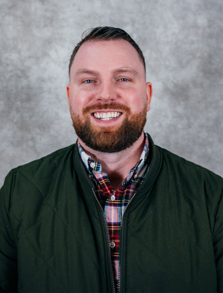 A man with a beard and plaid shirt smiles for a photo. He has a short haircut and wears a dark green jacket.