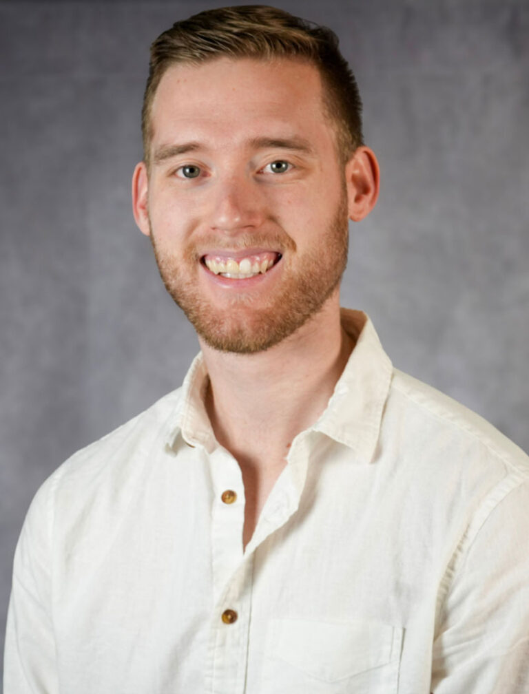 Young man with short brown hair and a short beard smiling at the camera. He is wearing a white button-up shirt.