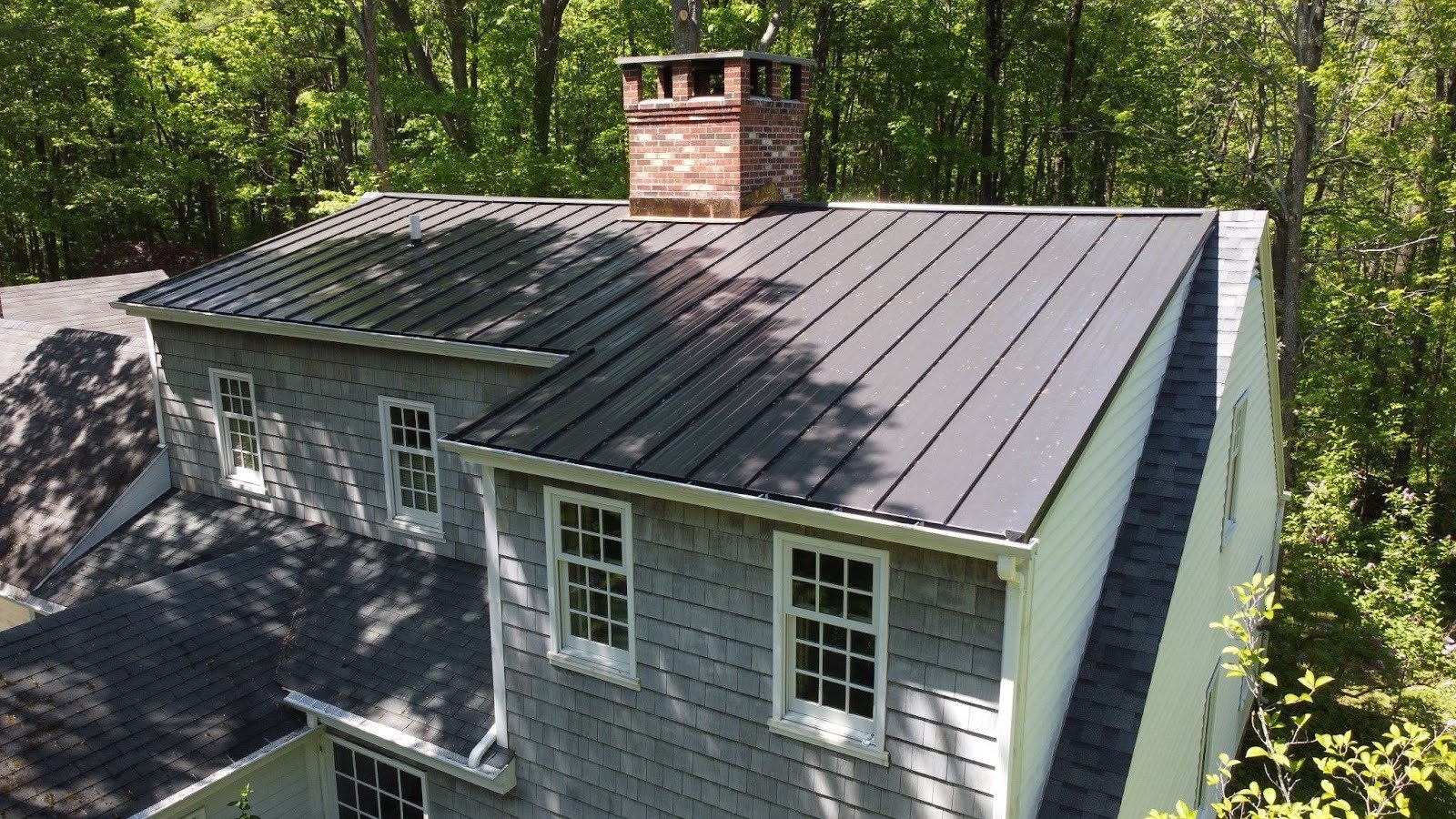 Gray house with metal roof and brick chimney. The house has white trim and is surrounded by trees.