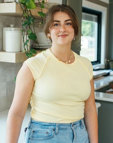 Young woman smiling in a kitchen with a green plant. She is wearing a yellow shirt and jeans.