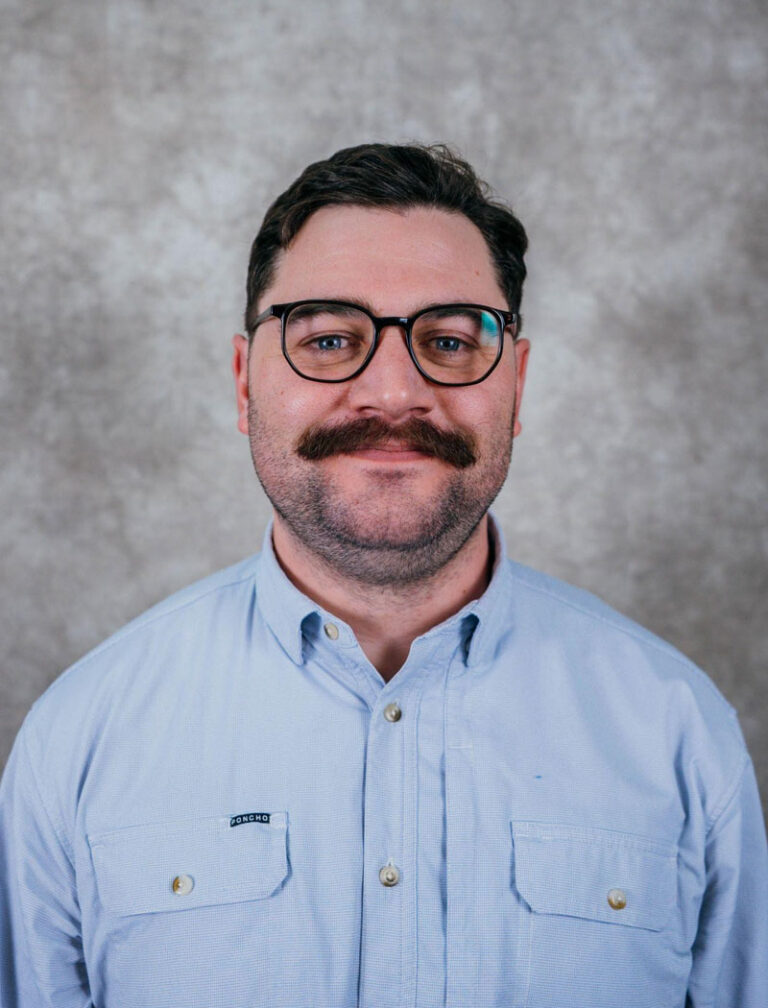 A man with glasses and a mustache wearing a blue shirt. He is looking directly at the camera.