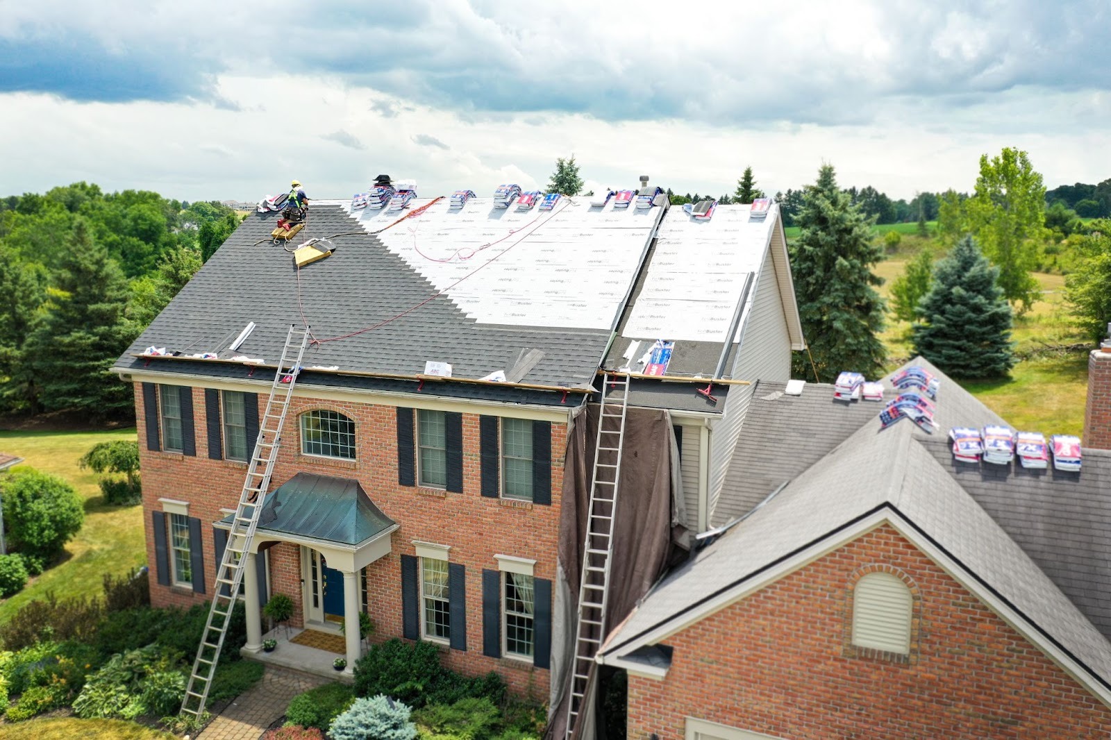 Workers on roof installing new shingles. Large house with brick exterior.