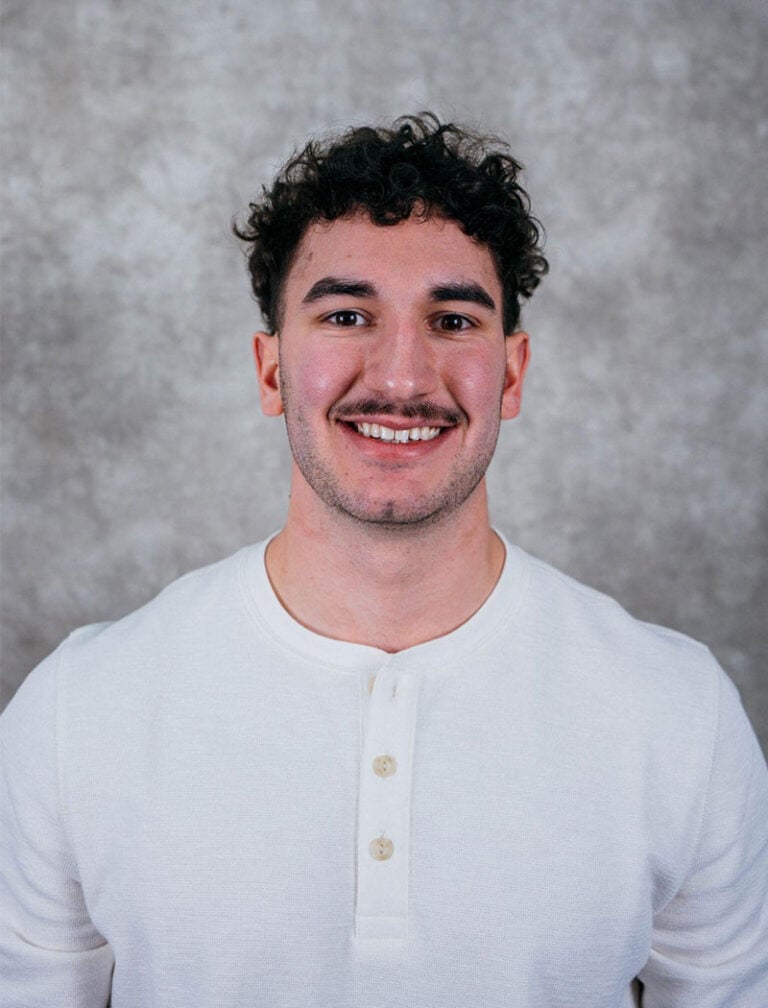 A young man with curly hair wearing a white shirt. He is smiling at the camera.