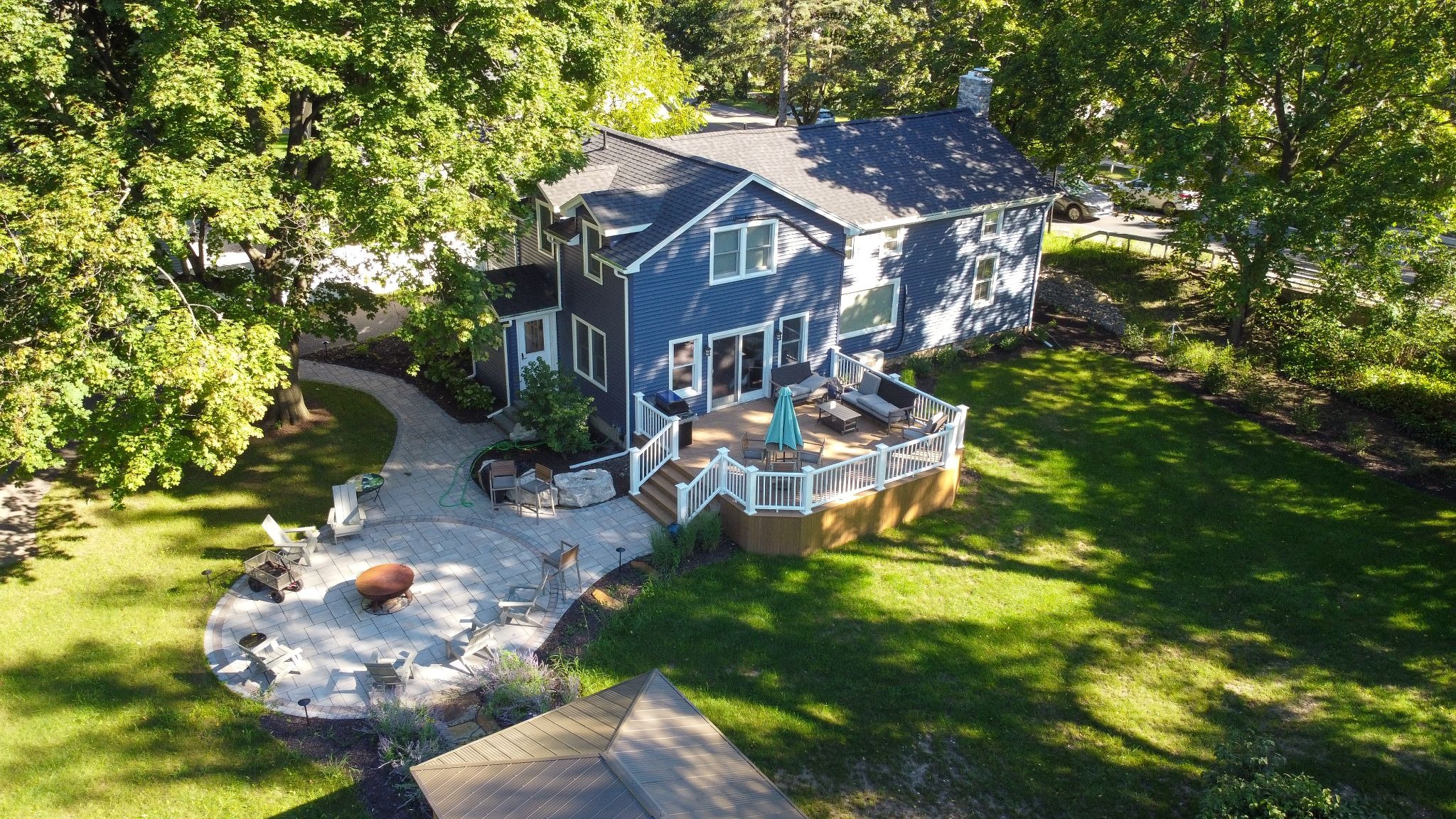 Aerial view of a large blue house with a deck and patio area. The house has a fire pit surrounded by stones.