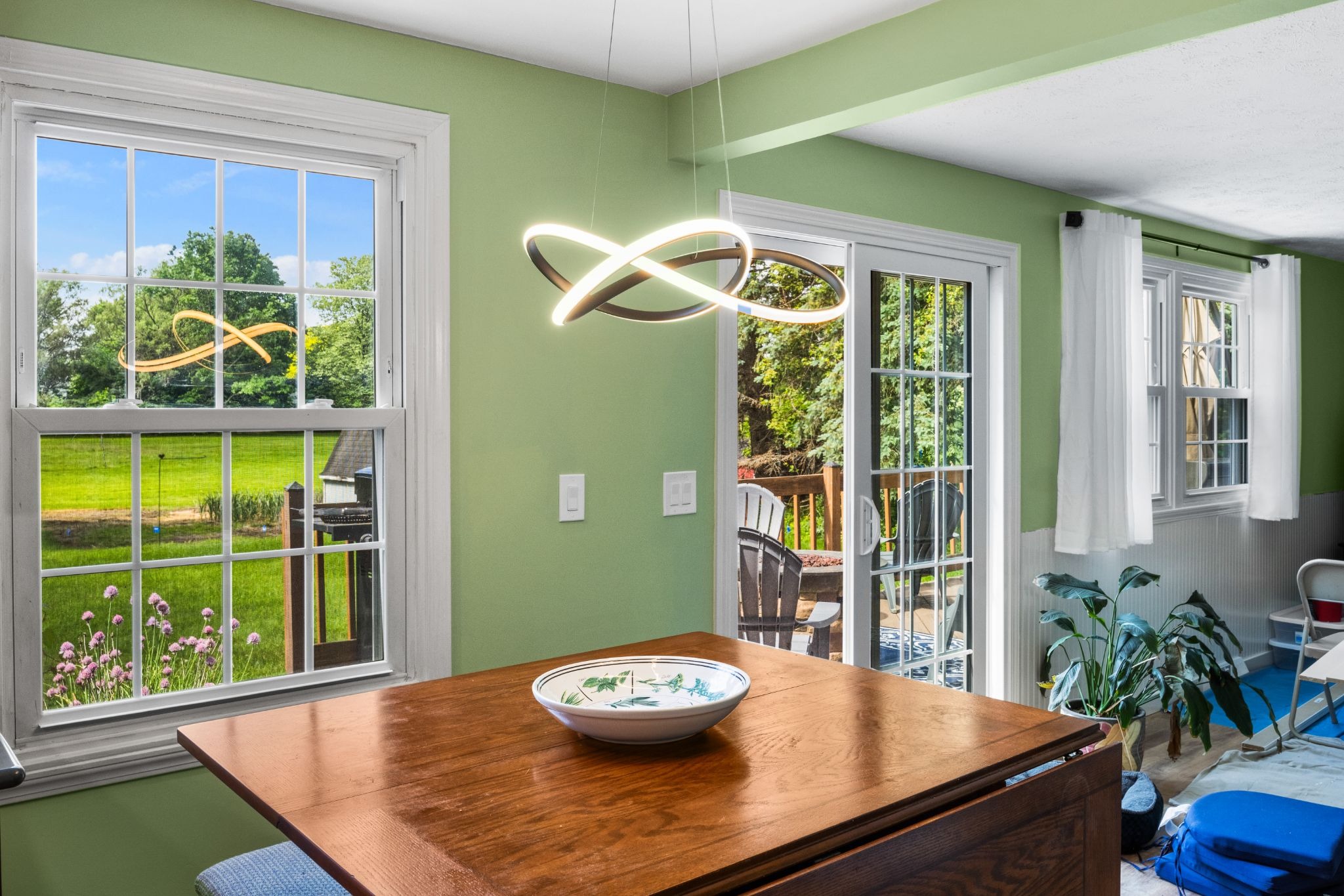A dining room with green walls, a wooden table, and a bowl on top. The room has large windows and a sliding glass door.