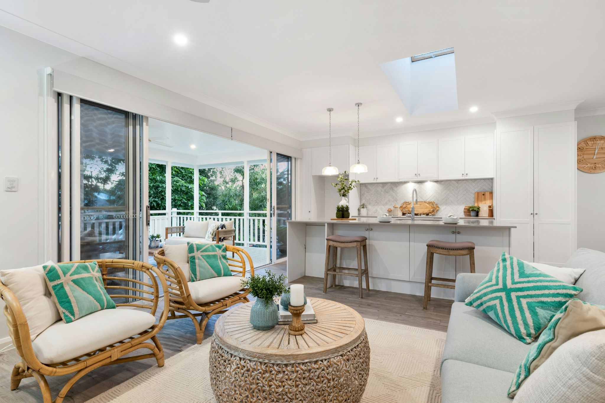 A modern living room with a kitchen in the background and a view of the outdoors through large windows. The room is furnished with a couch, coffee table, and chairs.