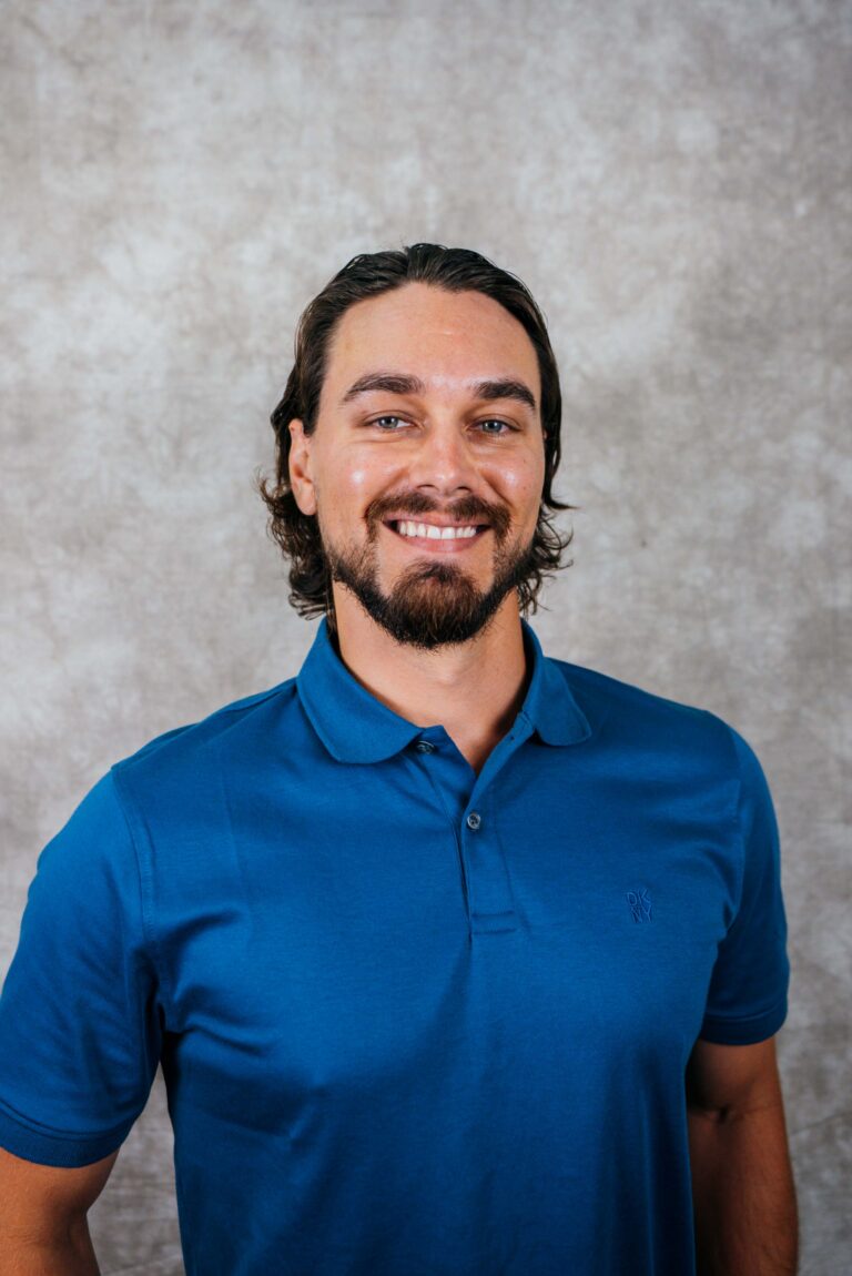 A smiling man with a beard and long hair, wearing a blue polo shirt, against a neutral background.