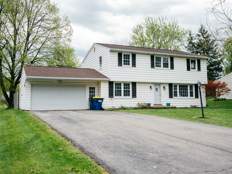 A white two-story house with black shutters, an attached garage, and a paved driveway. The front yard features green grass and trees.