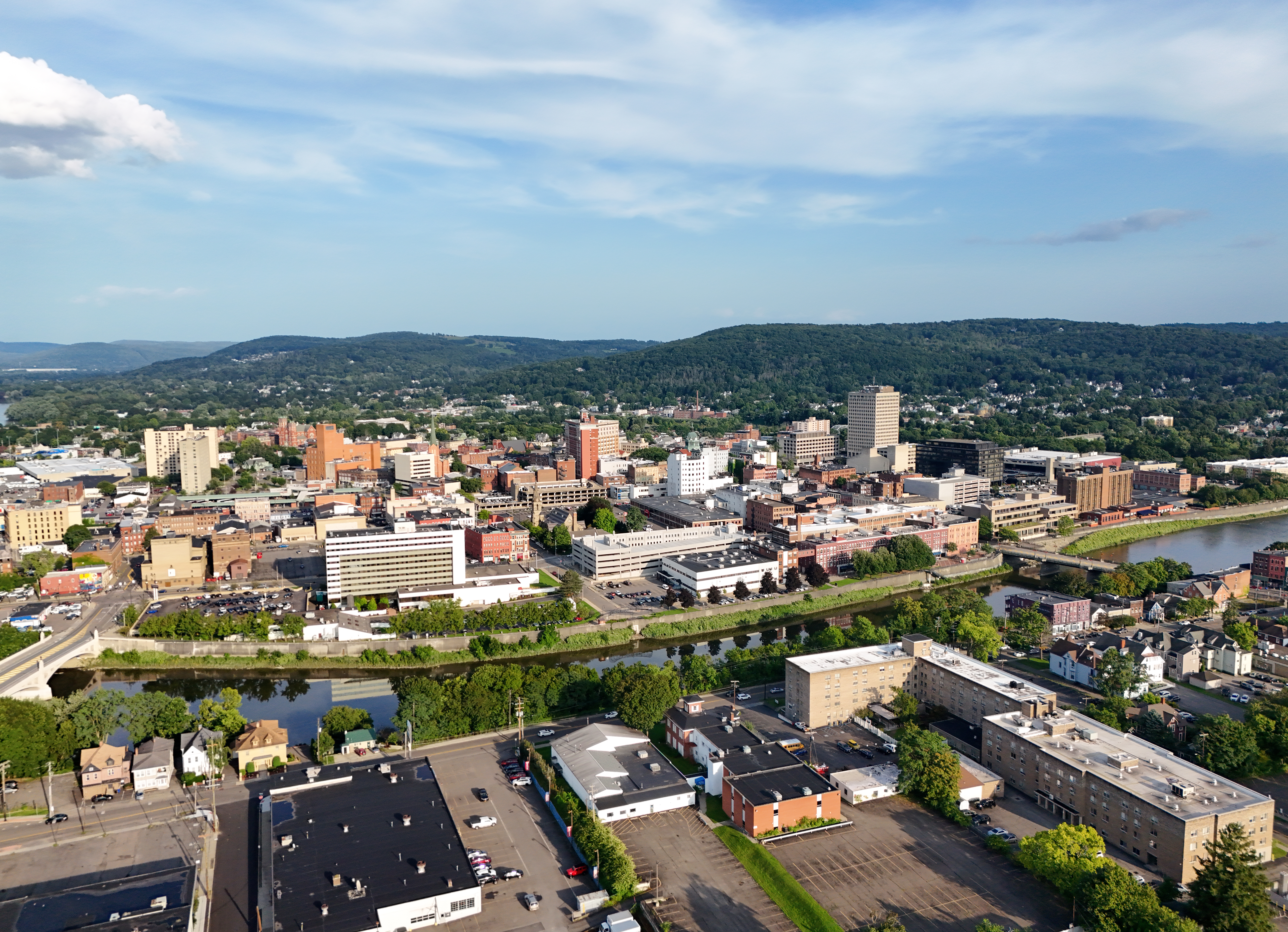 A cityscape viewed from above, with a winding river, dense brick buildings, and rolling green hills rising in the distance.