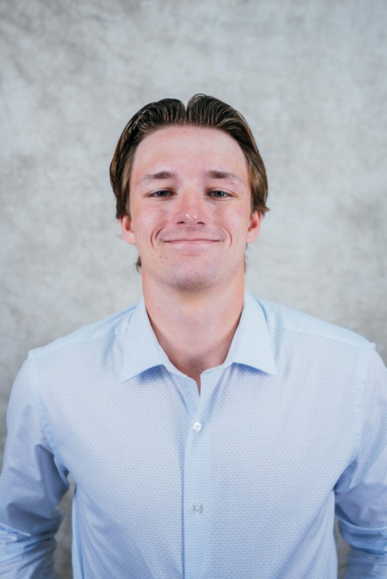 A smiling man with light brown hair wearing a light blue button-down shirt against a gray marbled background.