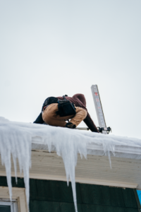 A worker on a snowy roof edge uses a ladder and safety harness to manage icicles.