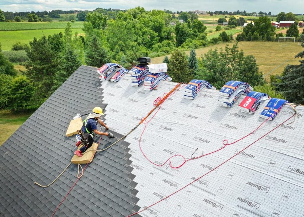 A worker installing grey shingles on a house roof overlooking green farmland, surrounded by stacks of roofing materials.