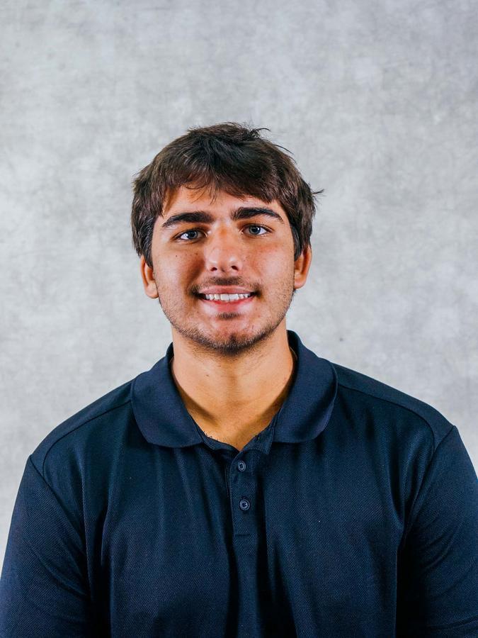 Portrait of a smiling young man wearing a dark blue polo shirt against a neutral gray background.