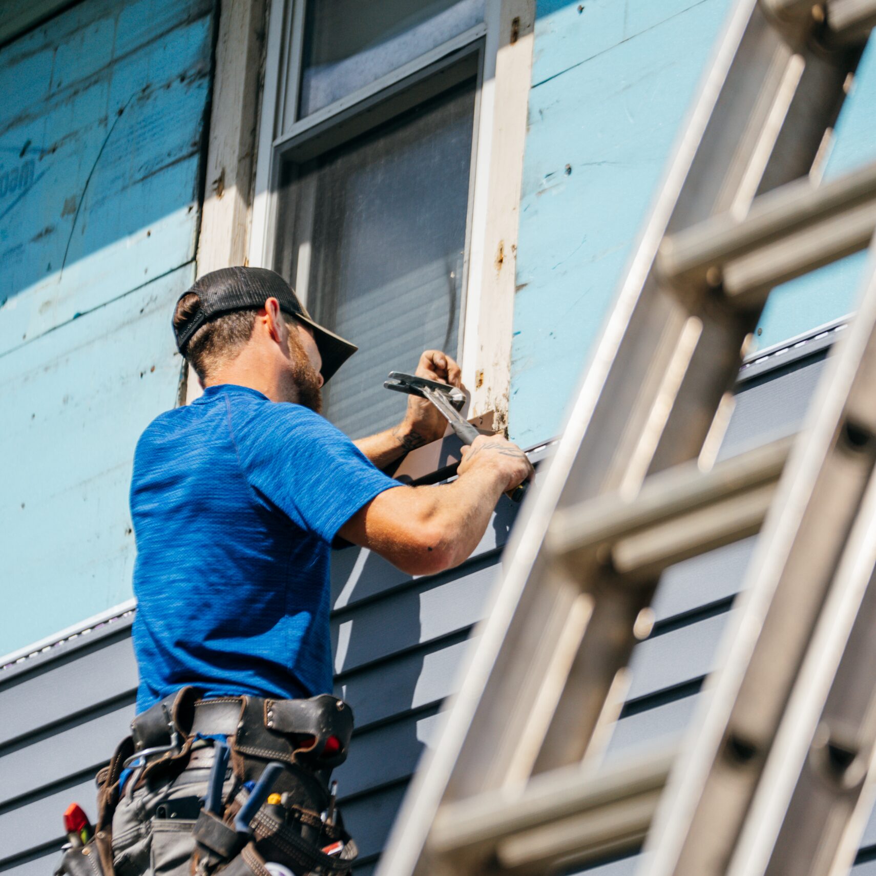A worker in a blue shirt and tool belt uses a hammer to install siding on a house while standing on a ladder, with more images below.