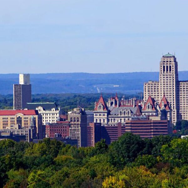 A cityscape with tall buildings and trees in the foreground. The background features a clear blue sky.