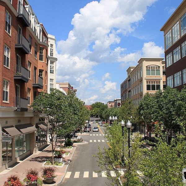 Urban street with multi-story buildings and green trees. A blue sky with clouds is visible in the background.