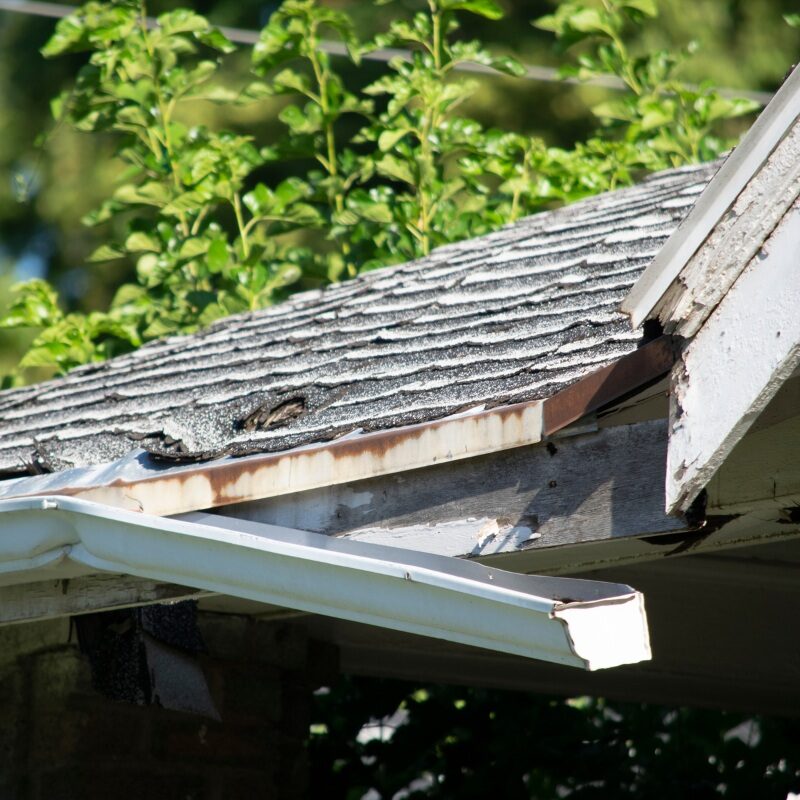 A close-up of a roof's edge with damaged gutters and missing shingles. The roof appears to be in disrepair.