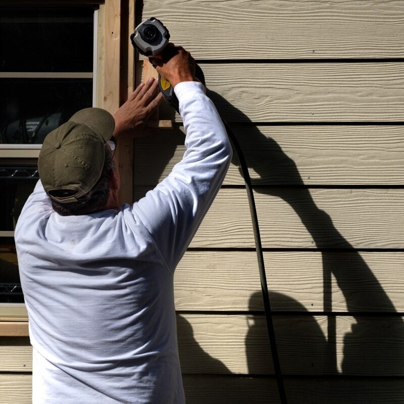 Man in a hat drilling into house siding. He is using a power tool.