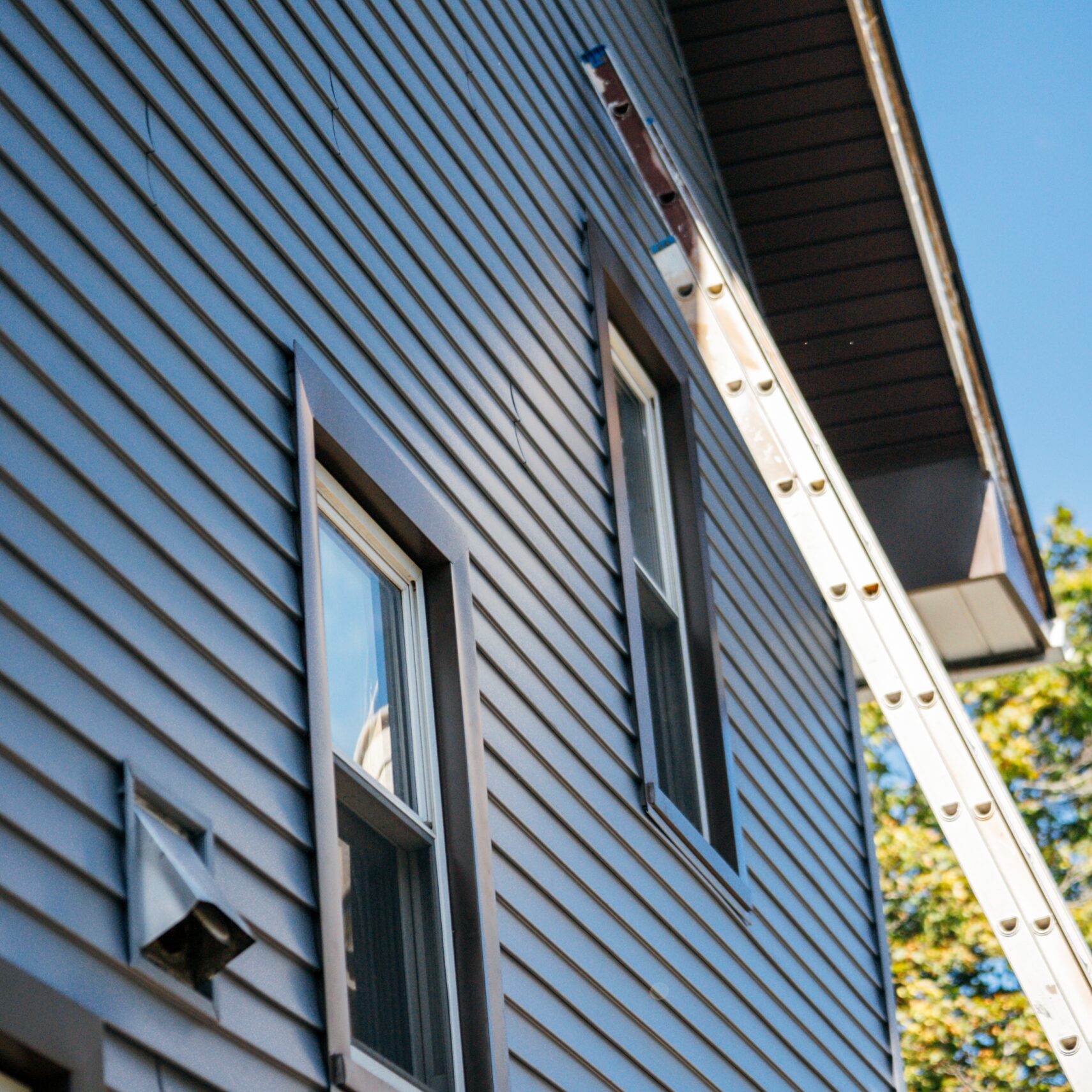 Ladder leaning against a blue-sided house with clear sky and green trees in the background.