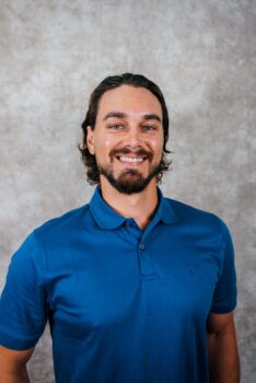 A smiling man with a beard and long hair, wearing a blue polo shirt, against a neutral background.