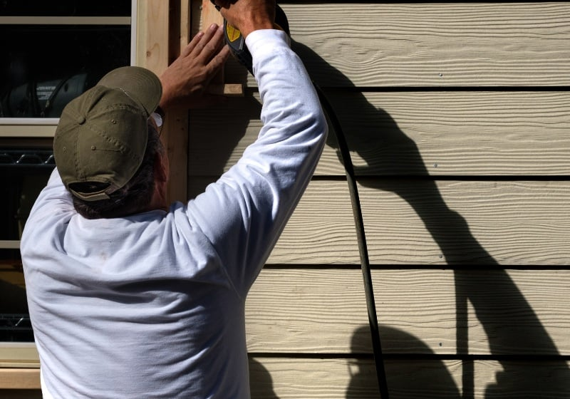 Man in a hat drilling into house siding. He is using a power tool.